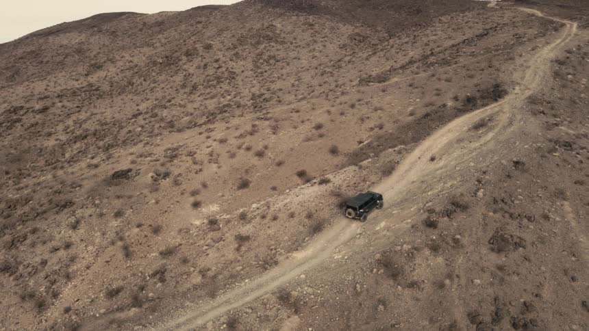 The video features a dark suv driving along a rugged dirt trail carved into a steep desert hillsideas the camera pulls back it reveals a vast arid landscape characterized by rolling hills and distant mountain ranges under a clear sky