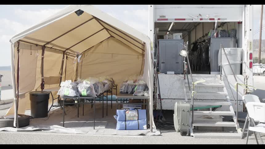 A tan canopy tent and the open back of a white equipment truck stand on an asphalt lot with hills in the background