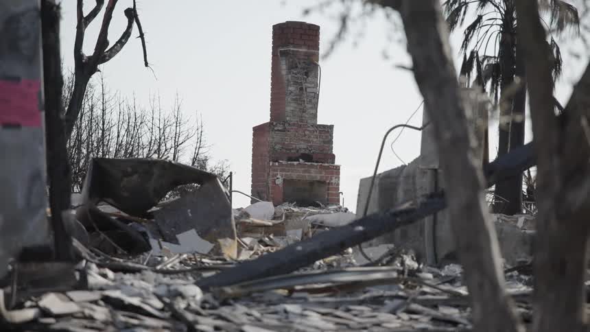 A brick chimney stands alone amidst the smoldering remains of a destroyed home where piles of charred wood and metal debris cover the ground under a hazy sky