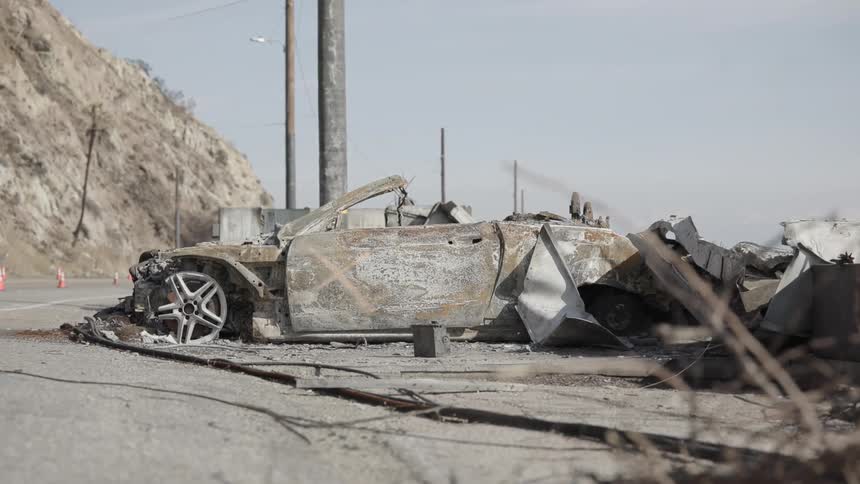 The video shows a stationary wide shot of a luxury convertible that has been completely gutted by fire on the side of a road debris and scorched metal surround the wreckage against a backdrop of a dry hilly landscape