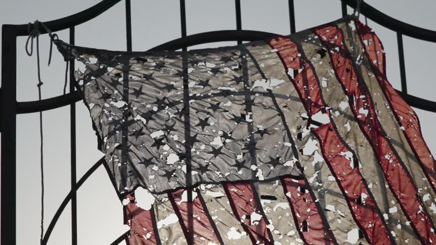 Decayed american flag full of holes hanging from a metal gate outdoors during day time