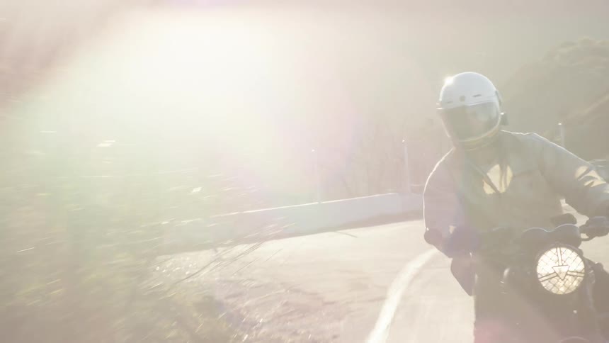 A man wearing a white helmet and tan jacket rides a black motorcycle along a winding mountain road during a sunny afternoon while a white car passes by him in the opposite direction