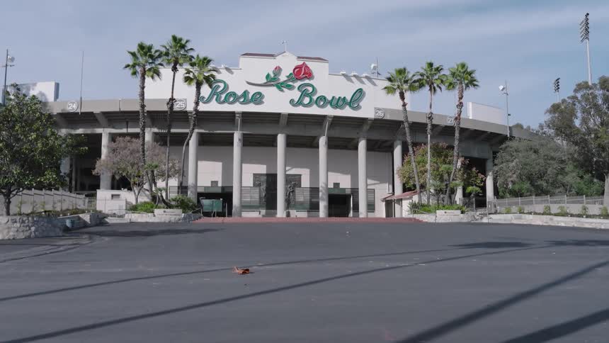 The video shows an exterior view of the iconic rose bowl stadium in pasadena california on a clear and sunny day the camera slowly zooms in toward the main entrance highlighting the famous logo and the surrounding palm trees
