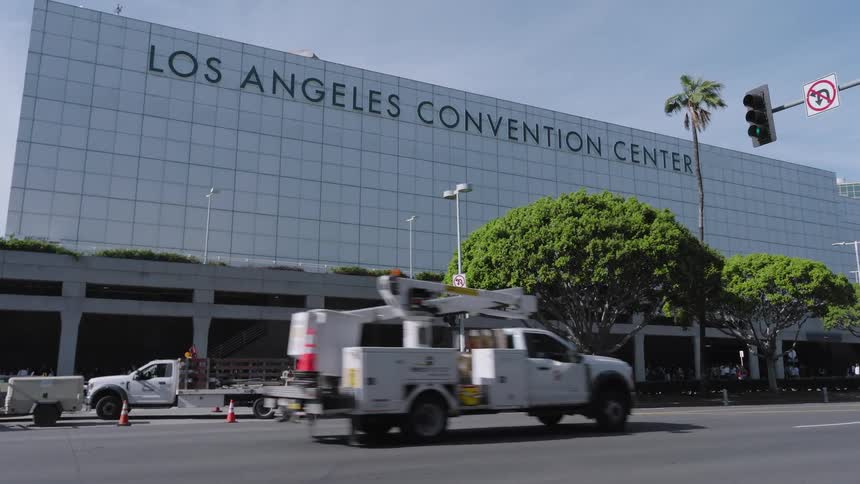 Cars and utility trucks drive past the los angeles convention center on a sunny day with green trees and palm trees lining the street under a clear blue sky
