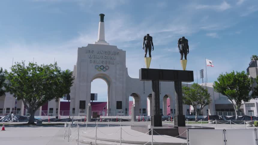 Los Angeles Memorial Coliseum entrance with two headless bronze statues standing on a black plinth under a clear blue sky surrounded by green trees and a paved courtyard with silver security chains