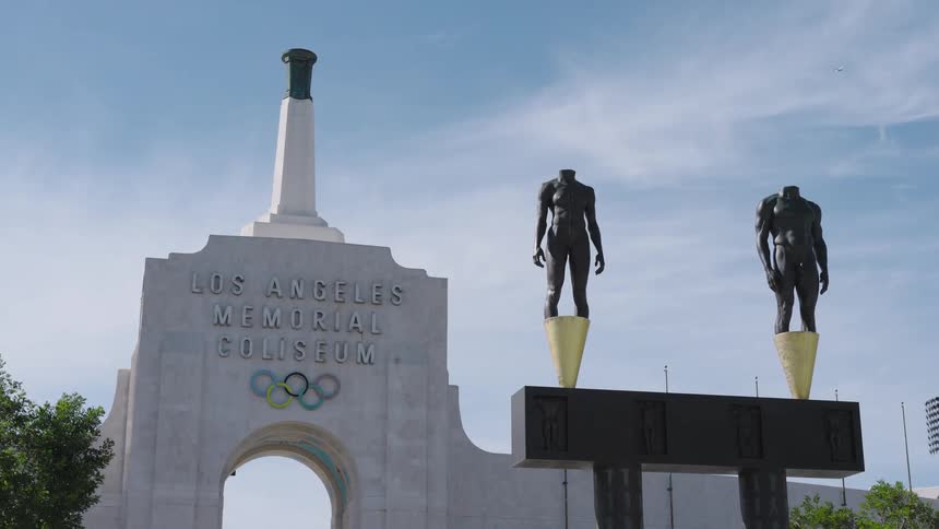 Entrance of the los angeles memorial coliseum with olympic rings and statues highlighted under a blue sky