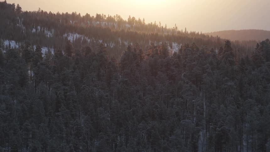 Snow on forested mountains as the sun glows in a hazy sky