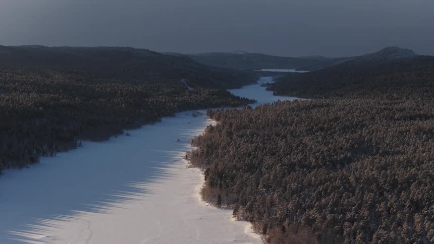 A wide frozen river winding through a dense pine forest covered in snow under an overcast sky