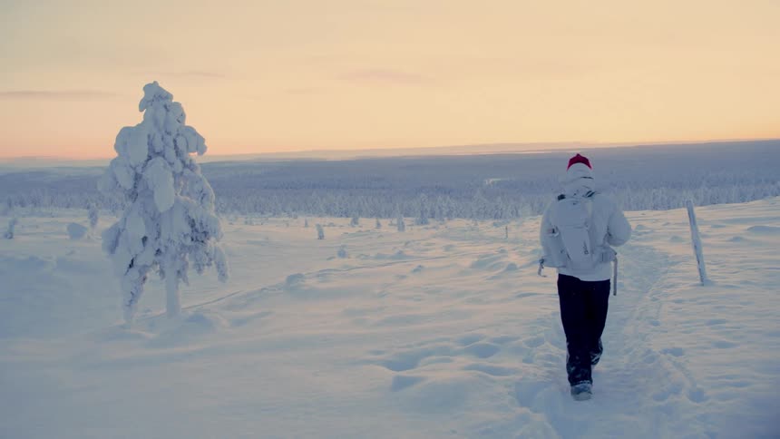 Person with backpack and red beanie walks across a snowcovered landscape at sunset