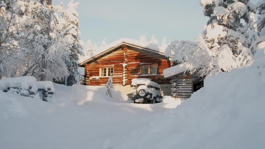 A wooden log cabin with a snow covered roof sits among frosted trees as golden sunlight hits the front of the house and a vehicle parked in the driveway