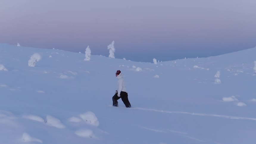 Adult man wearing a white coat and a beanie walking on a landscape fully covered in snow outdoors during dusk or dawn