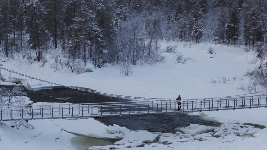 A person walks across a suspension bridge over a partially frozen river in a snowy forest the scene is a winter landscape with trees covered in snow and ice