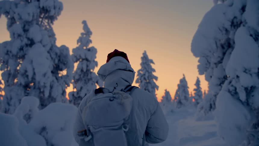 A person wearing a white winter coat and backpack walks through a snowy forest at sunset the sky is a warm orange and yellow contrasting with the cold snowcovered trees