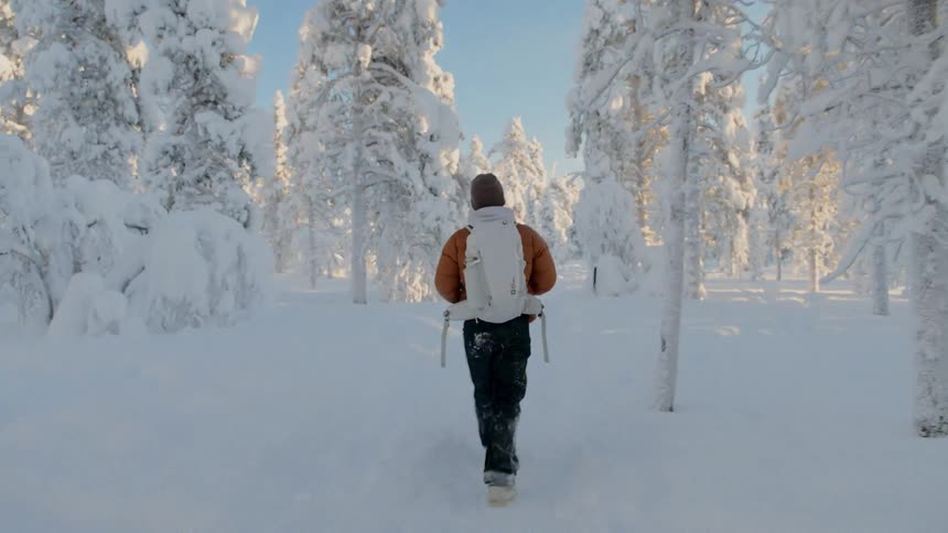 A man wearing an orange puffer jacket and a white backpack walks away from the camera through a deep snow covered forest during a bright winter day