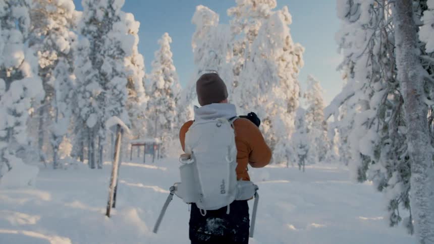 A man wearing an orange jacket and a white backpack walks through a thick snowy forest and raises a camera to take a photo of the frozen trees under a clear blue sky