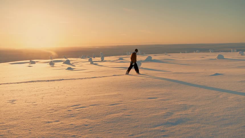 Person walks across a snowy plain casting a long shadow under golden sunset light