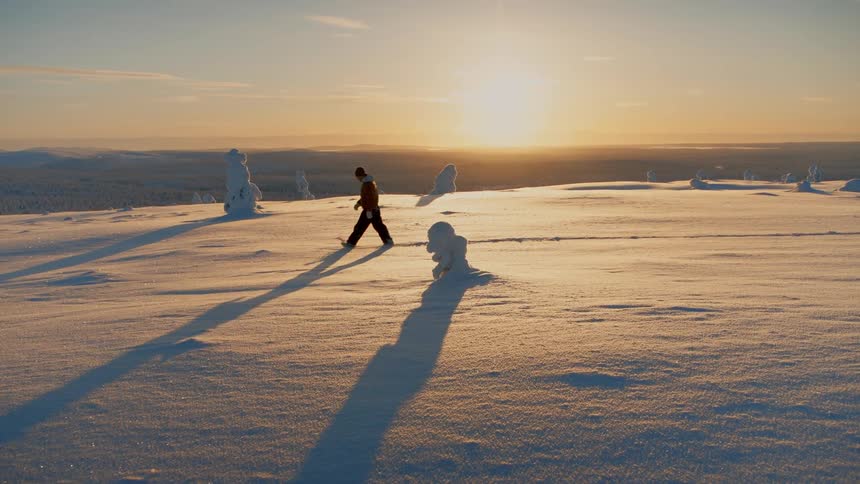 Person walking across snow covered land as the sun shines brightly on the horizon