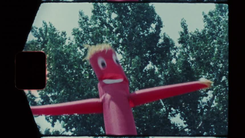 A red inflatable tube man with a yellow smiley face and thin hair moves and sways against a background of green leafy trees under a bright sky