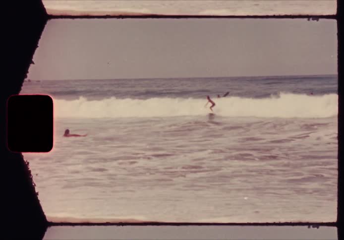 A person rides a wave on a surfboard as others swim in the ocean near a tropical shore in a vintage film style with a visible border