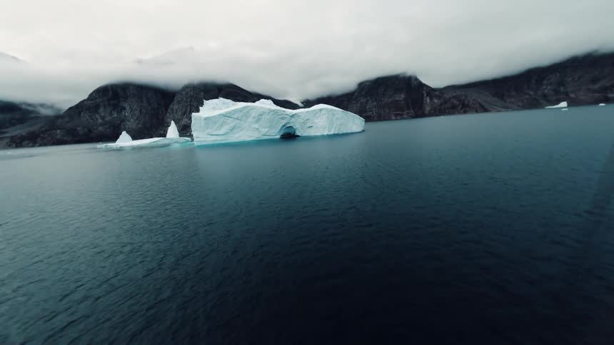 Sailing ship navigates in cold waters with an iceberg and mountains in the background