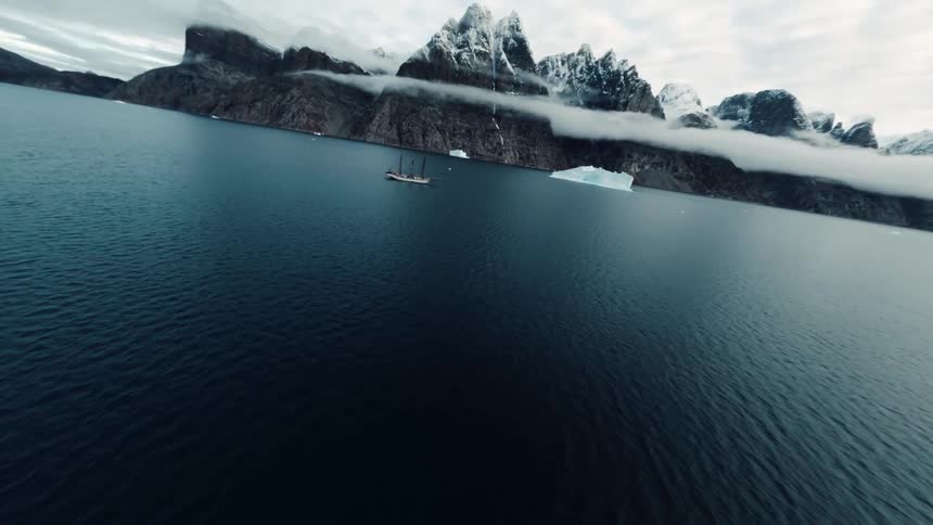 Sailboat sails on dark sea with snowcovered mountains and an iceberg in the background under a cloudy sky