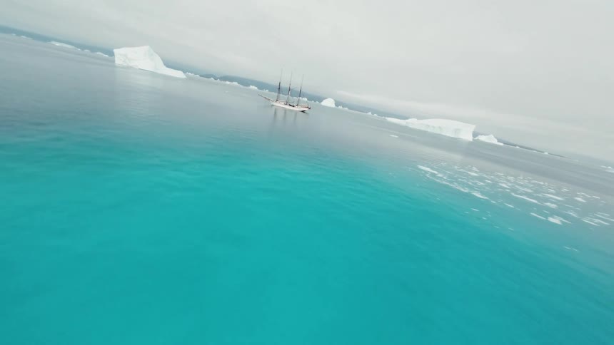 Sailing yacht floats in open water between icebergs on an overcast day