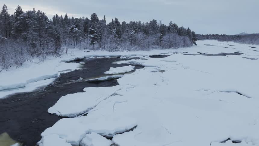 Broken ice on the river as it flows with a forest of snow covered trees along the coast
