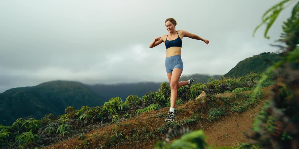 A young woman in athletic wear runs along a narrow dirt path on a lush green mountain ridge under a cloudy sky