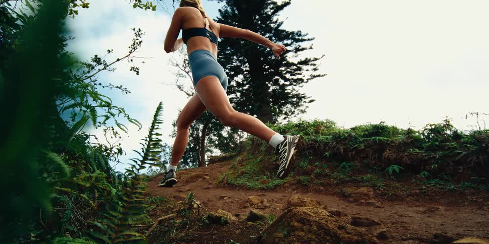 A woman wearing a dark sports bra and blue shorts runs up a steep dirt path surrounded by green ferns and trees under a bright sky