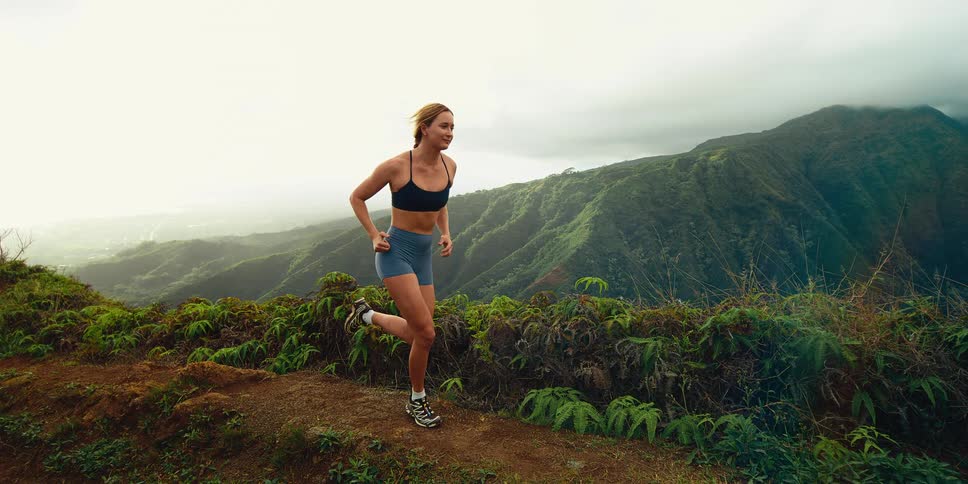 A blonde woman in a black sports bra and blue shorts runs along a narrow dirt trail on a lush mountain ridge overlooking a misty tropical valley under a cloudy sky