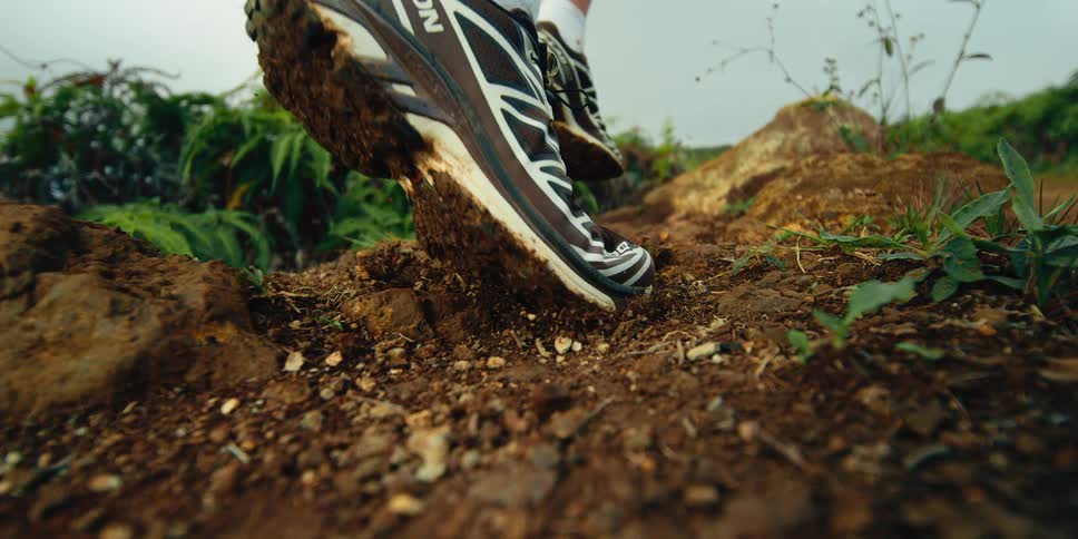A person wearing sneakers runs on a rugged dirt trail where the camera captures a close up of the foot kicking up soil and pebbles as the person moves away into a lush green landscape