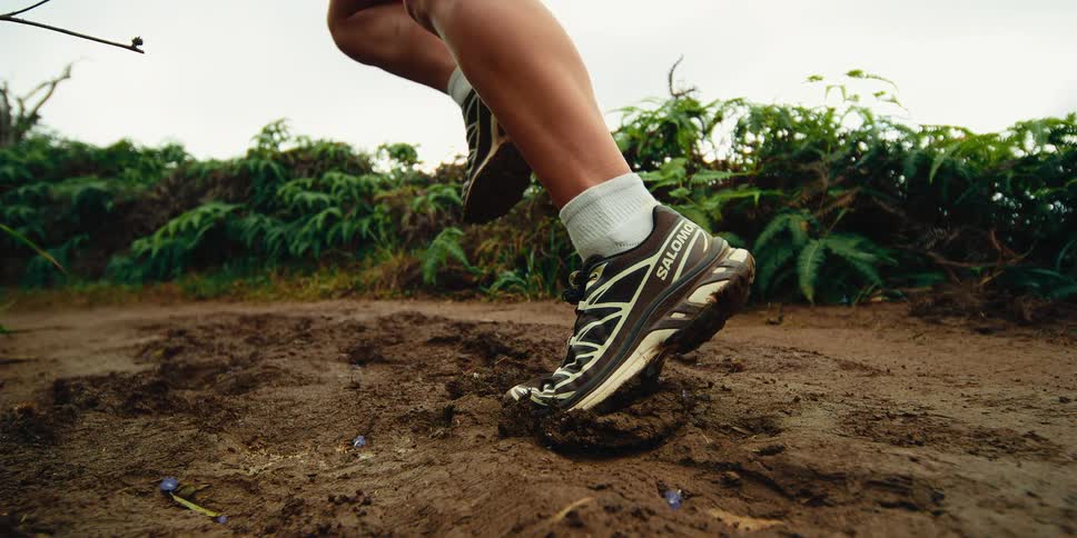 A person wearing salomon trail running shoes sprints across a muddy leafstrewn paththe impact of their foot leaves a deep impression in the soft earth sending small clods of mud flying into the air