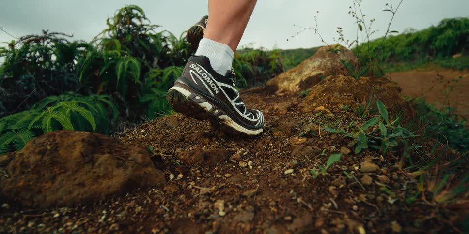 A close view shows the feet and legs of a woman wearing black running shoes and white socks as she runs along a dirt path with green plants and rocks