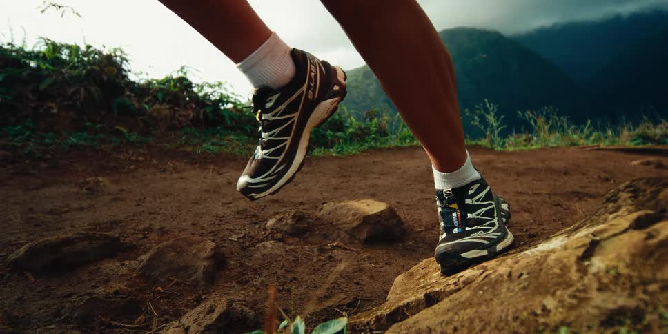A close view shows the feet of a woman wearing black running shoes and white socks as she jumps onto a large rock on a dirt path with green mountains in the distance