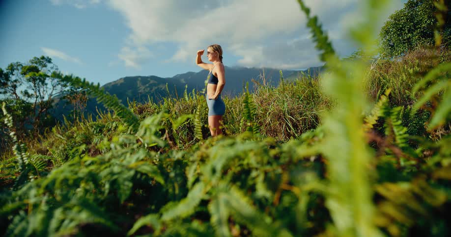 A woman wearing a dark sports bra and blue shorts stands on a grassy hill with her hand to her forehead while looking at distant mountains under a blue sky with soft white clouds