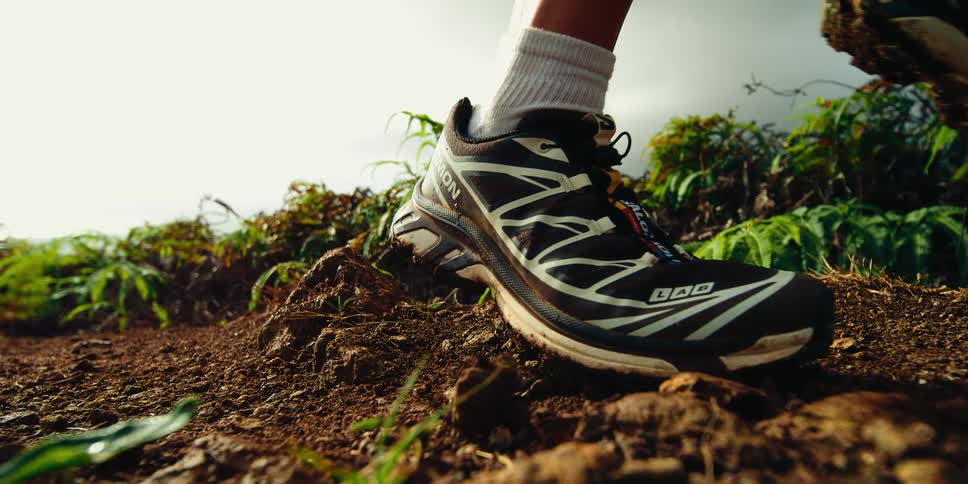 The video provides a lowangle slowmotion closeup of a person running across a dirt trail the impact of their athletic shoes kicks up loose soil and pebbles against a backdrop of lush green ferns