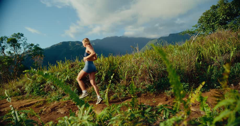 A woman wearing athletic gear runs across a dirt trail on a lush green ridge with a massive mountain range and cloudy sky in the background
