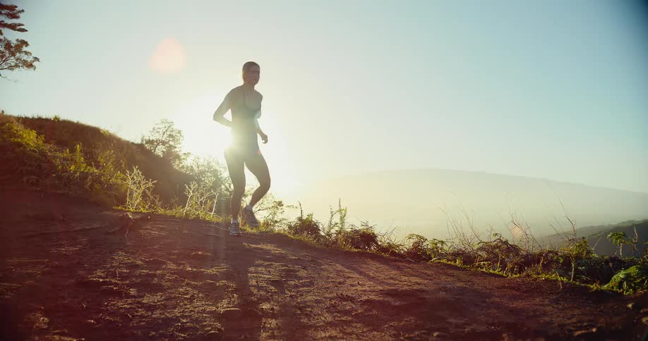 A blonde woman in athletic wear runs along a dirt mountain trail during the day