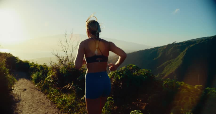 A woman running along a scenic mountain trail during a golden hour sunset she is captured from behind as she jogs toward the horizon overlooking a lush valley and distant peaks
