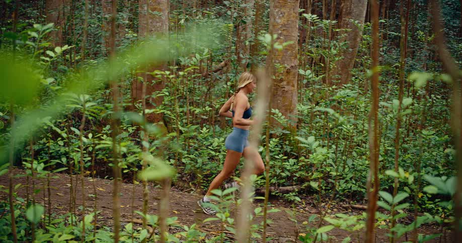 This video shows a woman running across a dirt path in a dense green forest she is captured in a sideprofile view as she moves through the tall trees and lush undergrowth