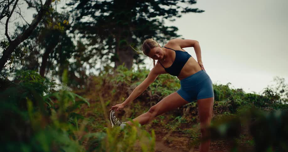 A woman in athletic wear performs a standing hamstring stretch on a forested trail she leans forward to touch her toes while keeping one leg elevated on the rising terrain