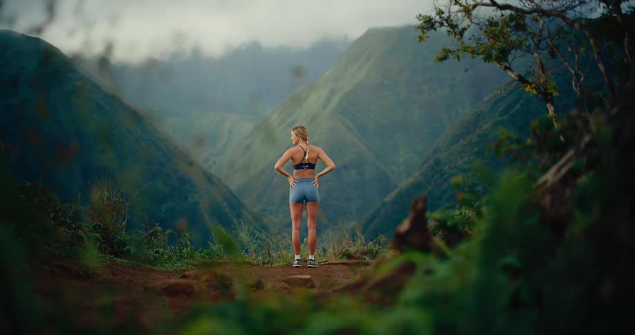 A woman wearing a dark sports bra and blue shorts runs up a dirt path and jumps over a large rock with green plants and mountains in the background under a blue sky