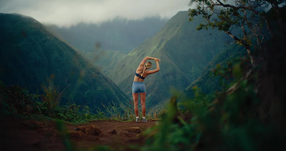 A blonde woman in athletic wear stands on a dirt trail with her back to the camera performing a triceps stretch while looking out over a vast green mountain valley under a misty sky