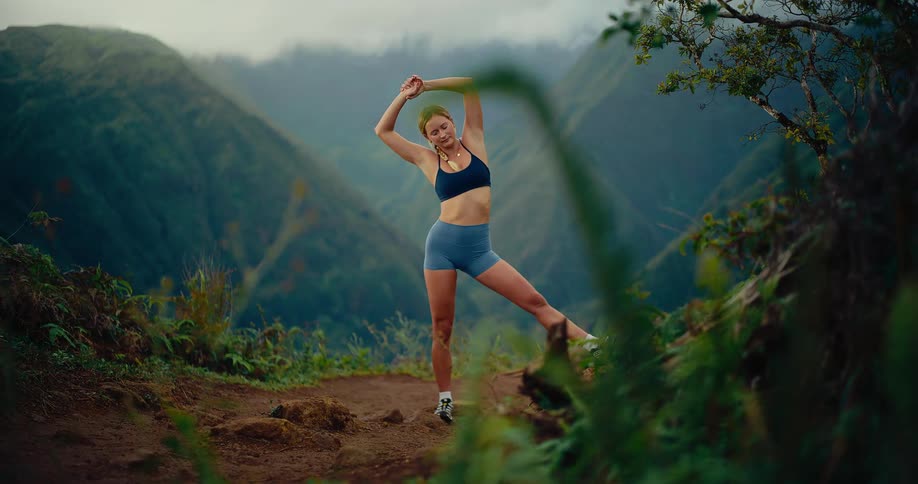 A woman wearing a dark sports bra and blue shorts stretches her legs and arms while standing on a dirt path overlooking a deep green valley with misty mountains in the background