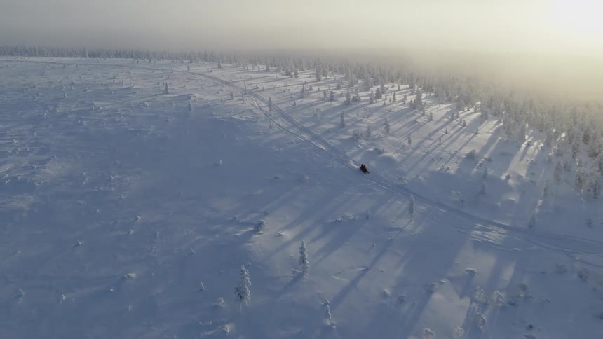 An aerial view shows a person driving a snowmobile through a snow covered forest during winter