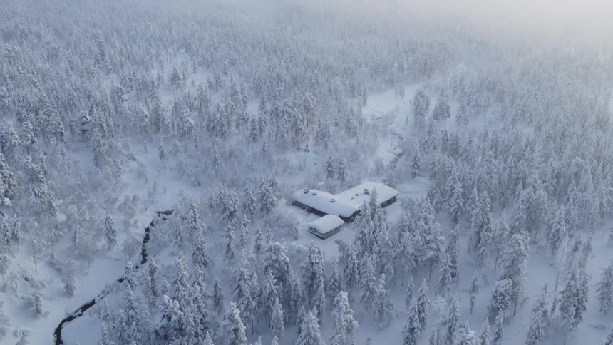 An aerial drone shot glides over a dense snowcovered forest where a dark winding stream cuts through the white landscape centered in the frozen wilderness is a large secluded building with snowpiled roofs appearing isolated amidst the misty frost