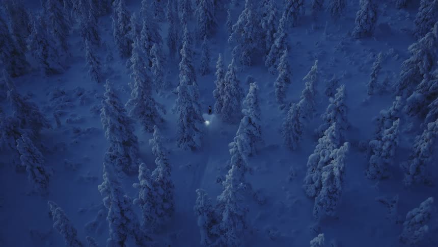 Aerial view of a person wearing a headlamp flashlight as they walk through a snow covered forest
