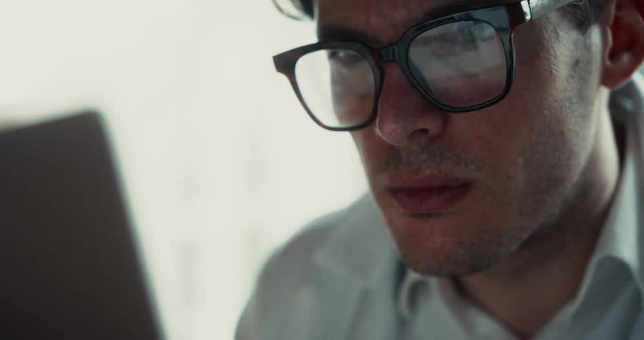 A man wearing glasses looks at a computer screen with reflections visible in his lenses