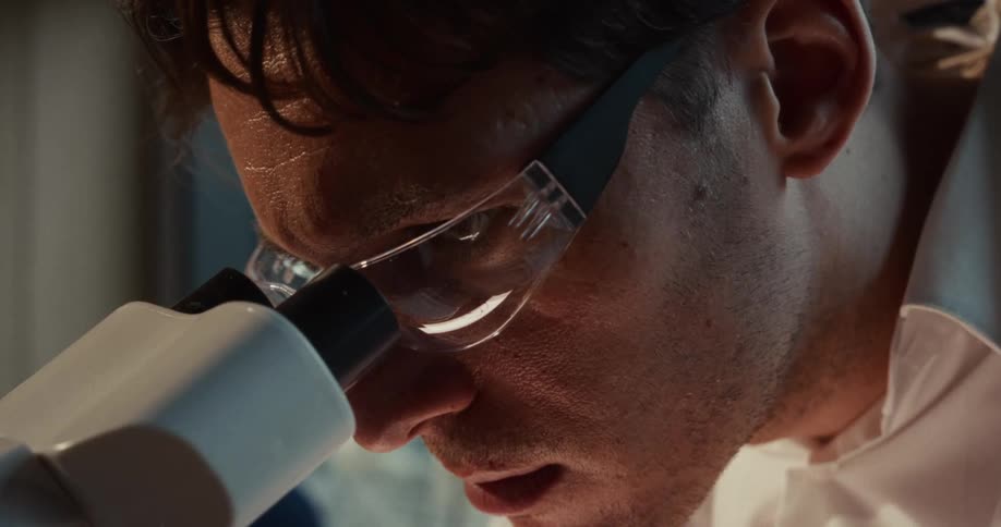 An up close view of a young man wearing googles looking in a microscope in a dimly lit room