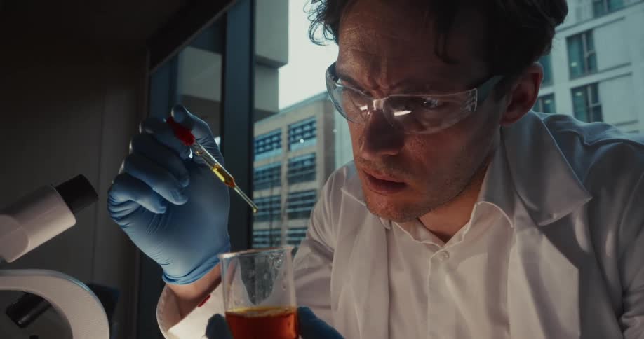 Male laboratory worker uses a dropper and a beaker in a lab with a view of office buildings during the daytime